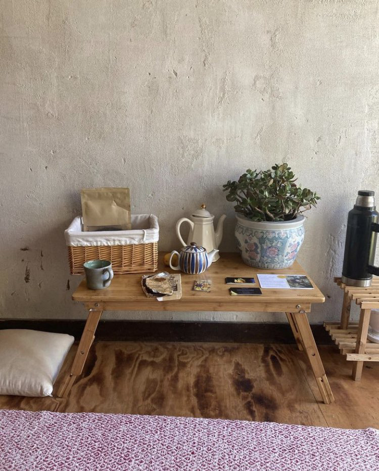 Small table with a jade plant, basket with tea bags in it, a tea cup, 2 ceramic kettles, and some fliers on top of it.